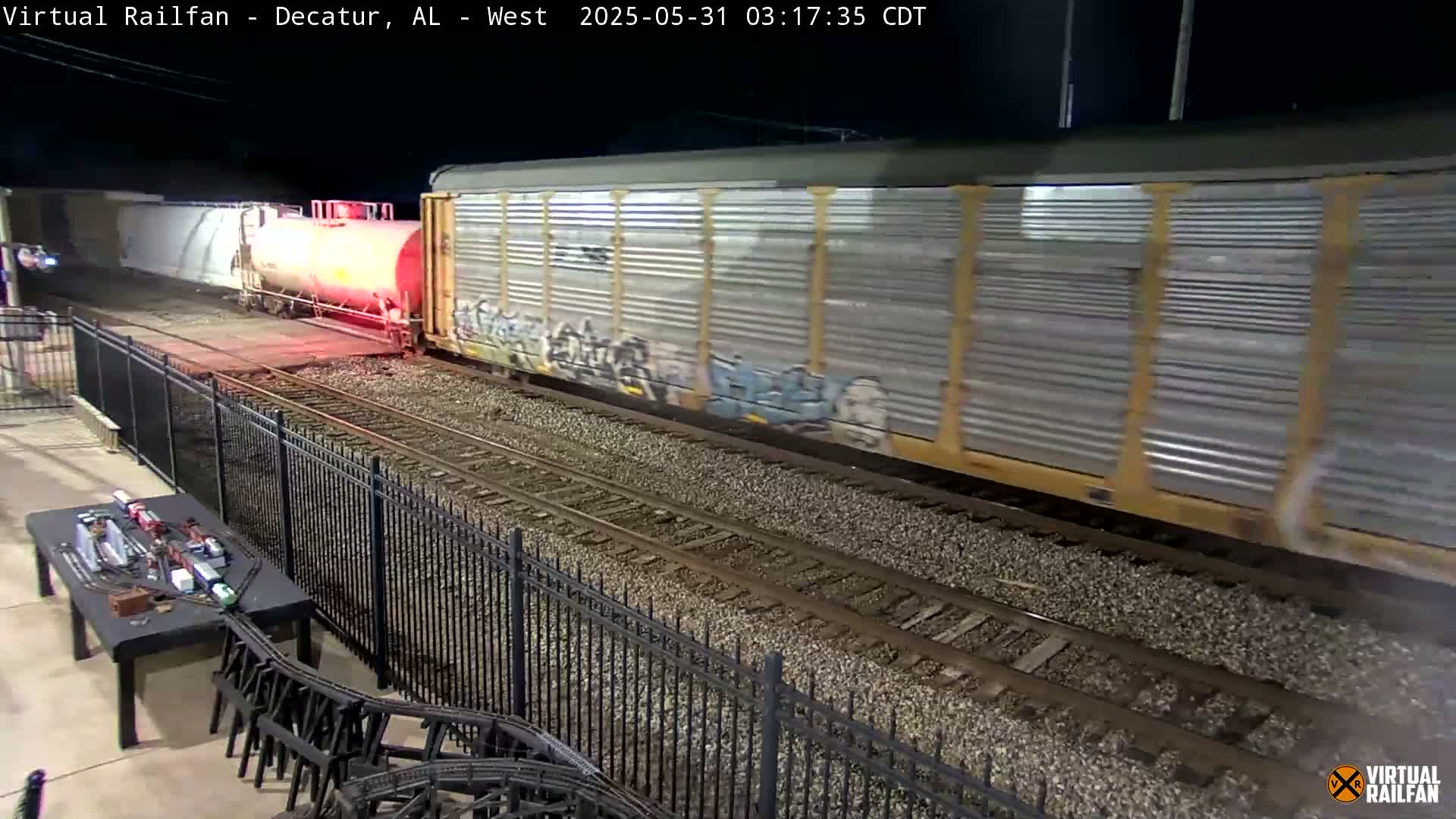 At night, a freight train with graffiti-covered cars passes by a black metal fence, next to a table with a model train set.