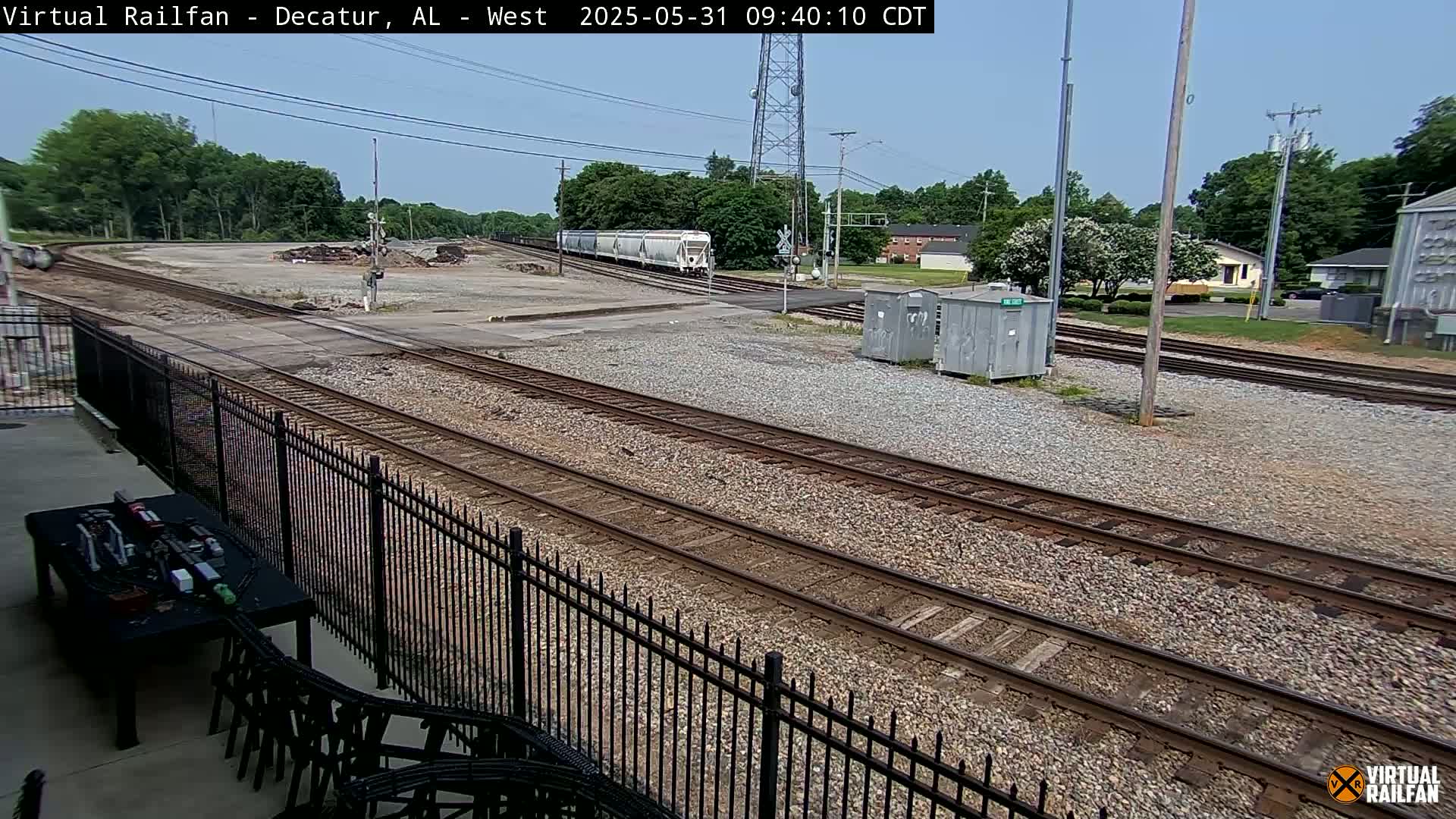 A long freight train passes through a rail yard on a sunny day, with several sets of tracks visible and a black metal fence in the foreground.