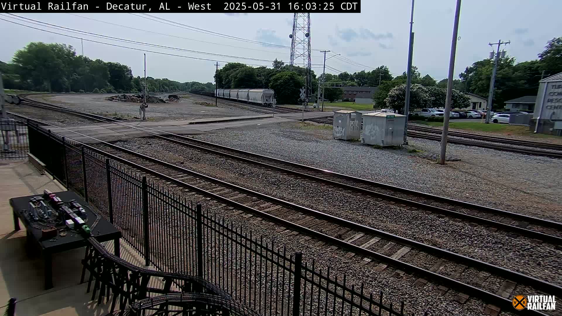 A partly sunny day shows several sets of train tracks converging near a fenced-in area with a model train set visible in the foreground.