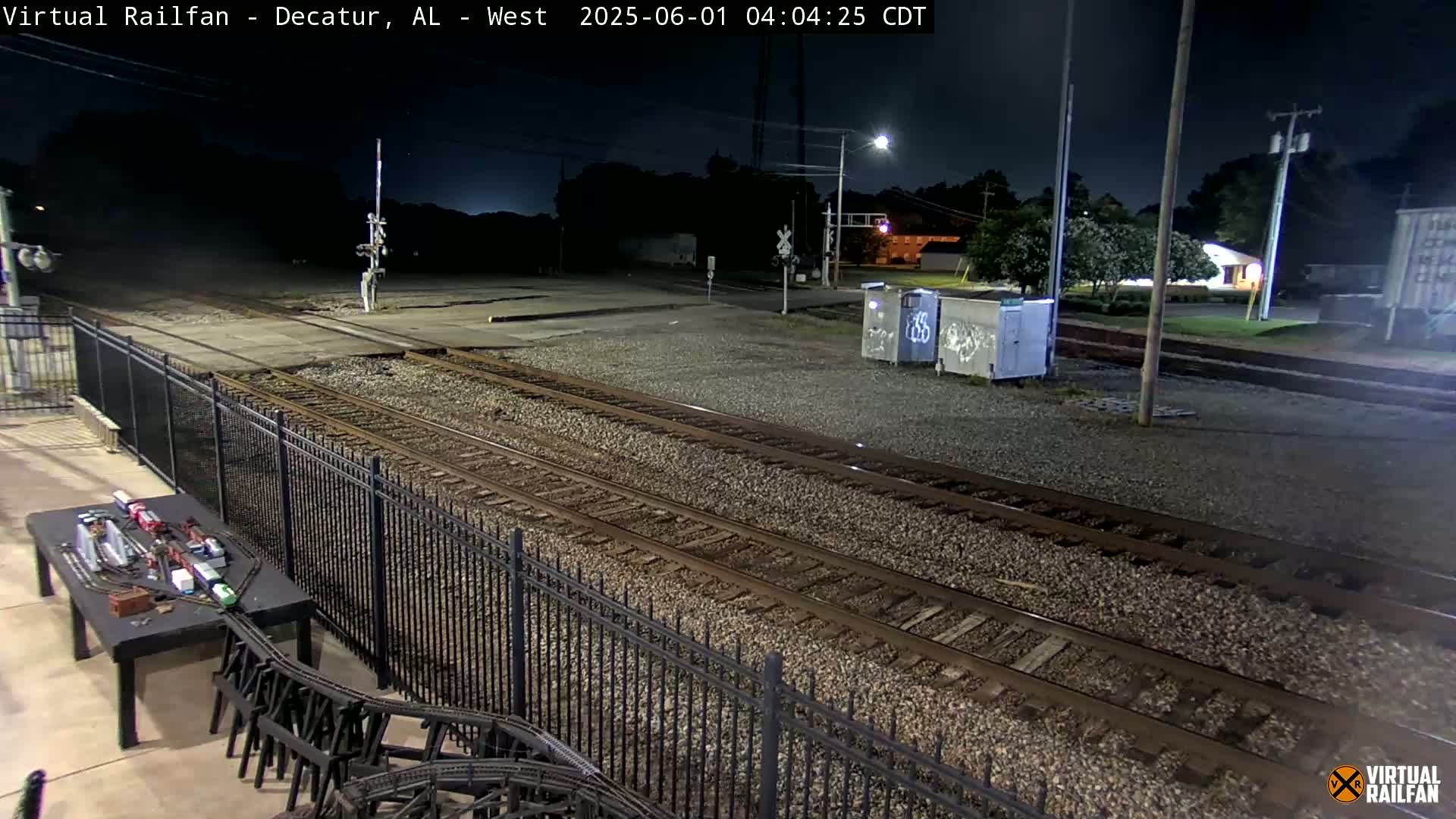 A nighttime view of a train yard with two sets of tracks, a black metal fence, and a model train set on a table in the foreground.