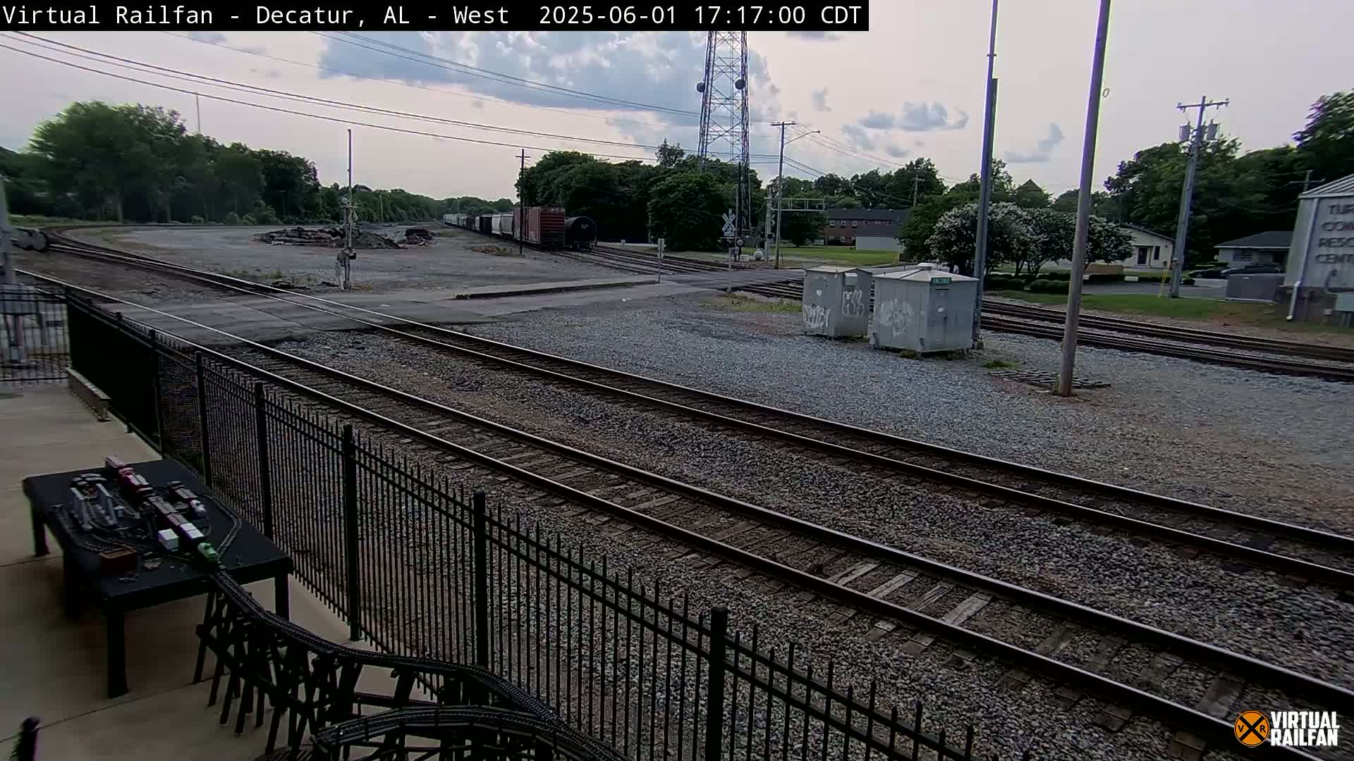 A railroad yard with several train tracks, a few boxcars, and some buildings in the background under a partly cloudy sky.