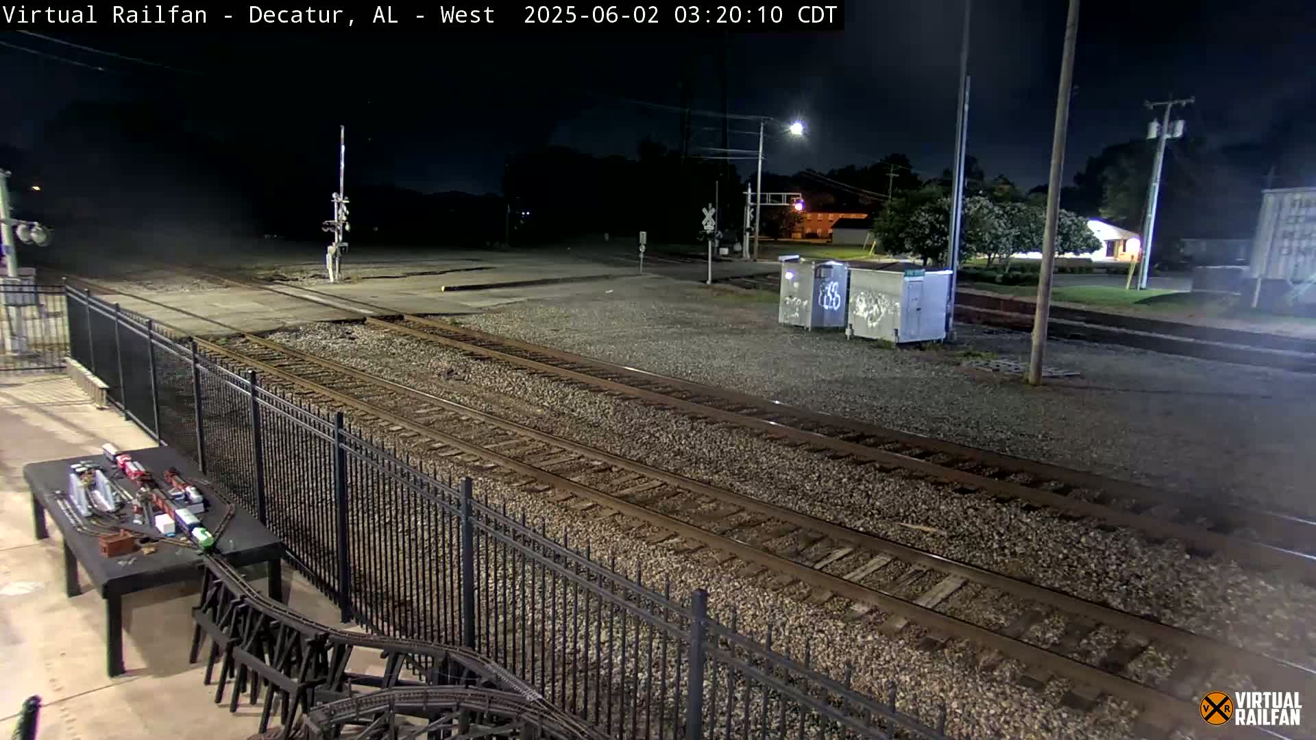 A nighttime view of train tracks, a metal fence, and a model train setup on a table.