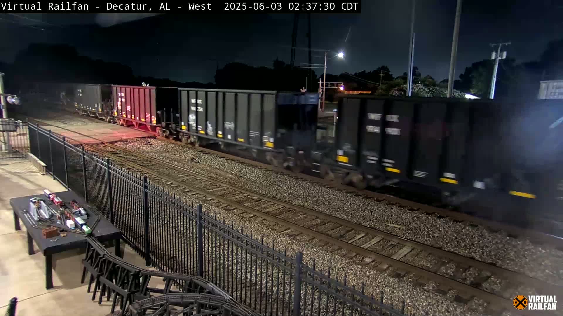 A long freight train moves along a track at night, next to a black metal fence and a model train set on a table.