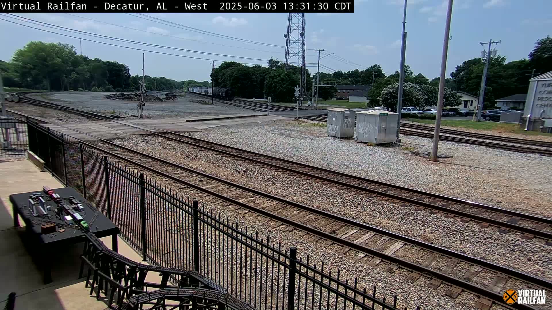 A sunny outdoor view of multiple train tracks, a gravel yard, and some nearby buildings, partially obscured by a black metal fence.