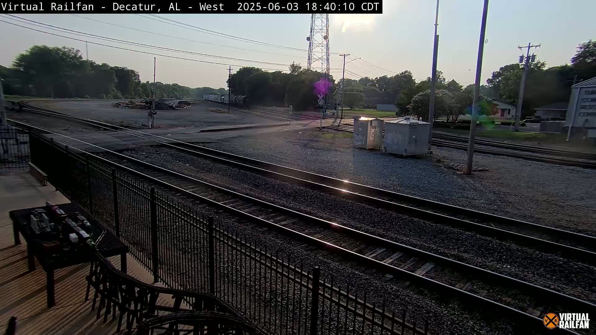 Several sets of train tracks wind through a gravel yard under a mostly clear, sunny sky, with some trees and buildings visible in the background.