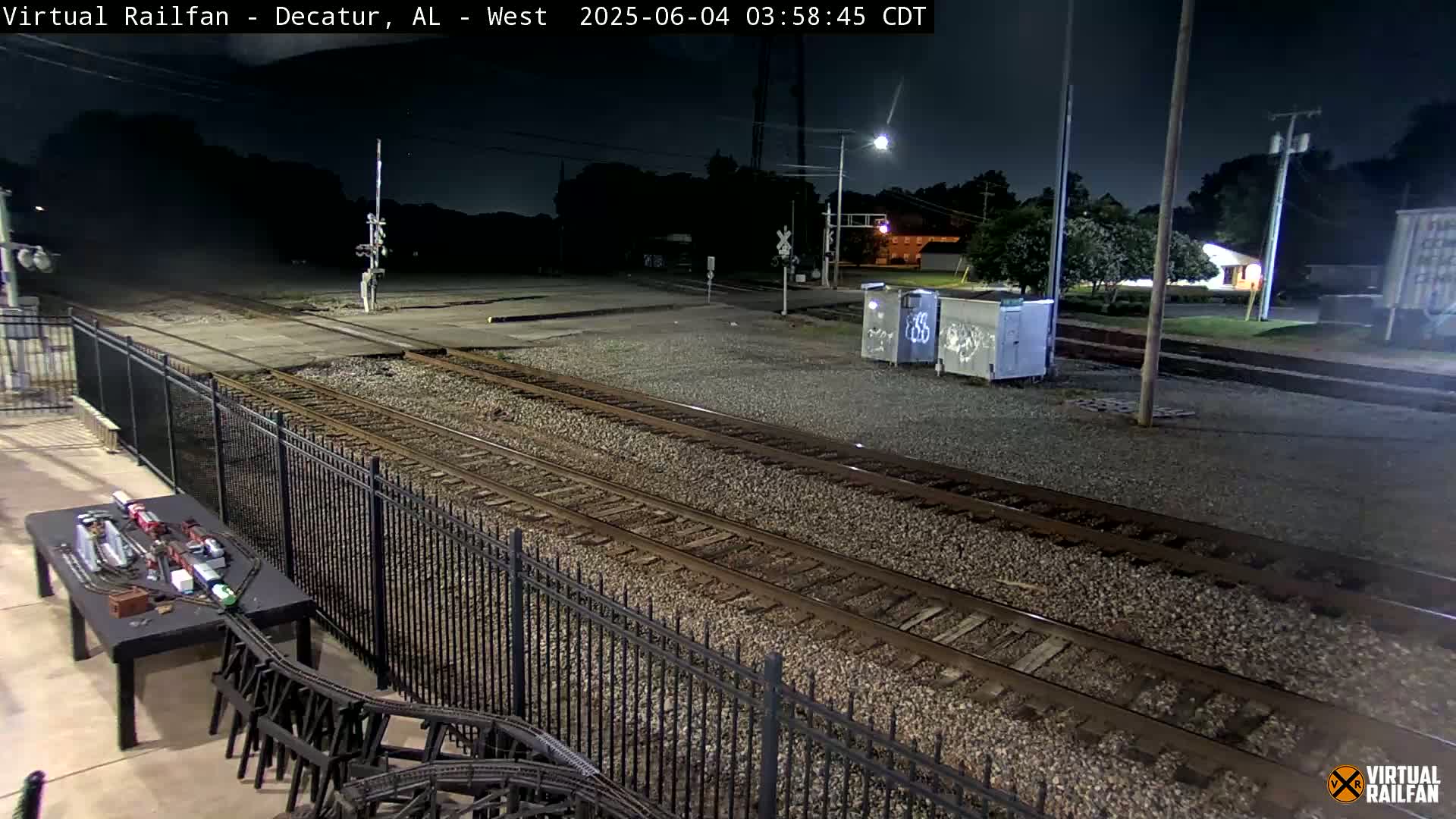 A nighttime view of railroad tracks and a model train set on a table, next to a black metal fence, under a dark, clear sky.