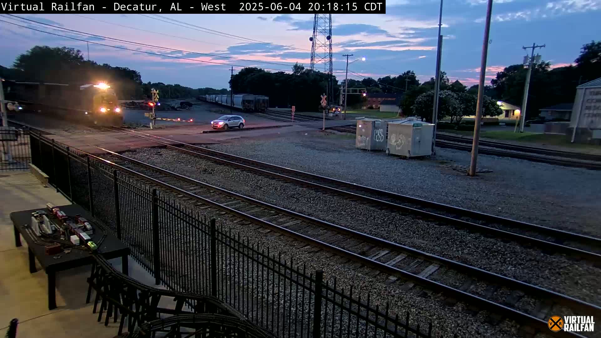 A train passes through a railroad crossing at twilight, with a car waiting nearby.