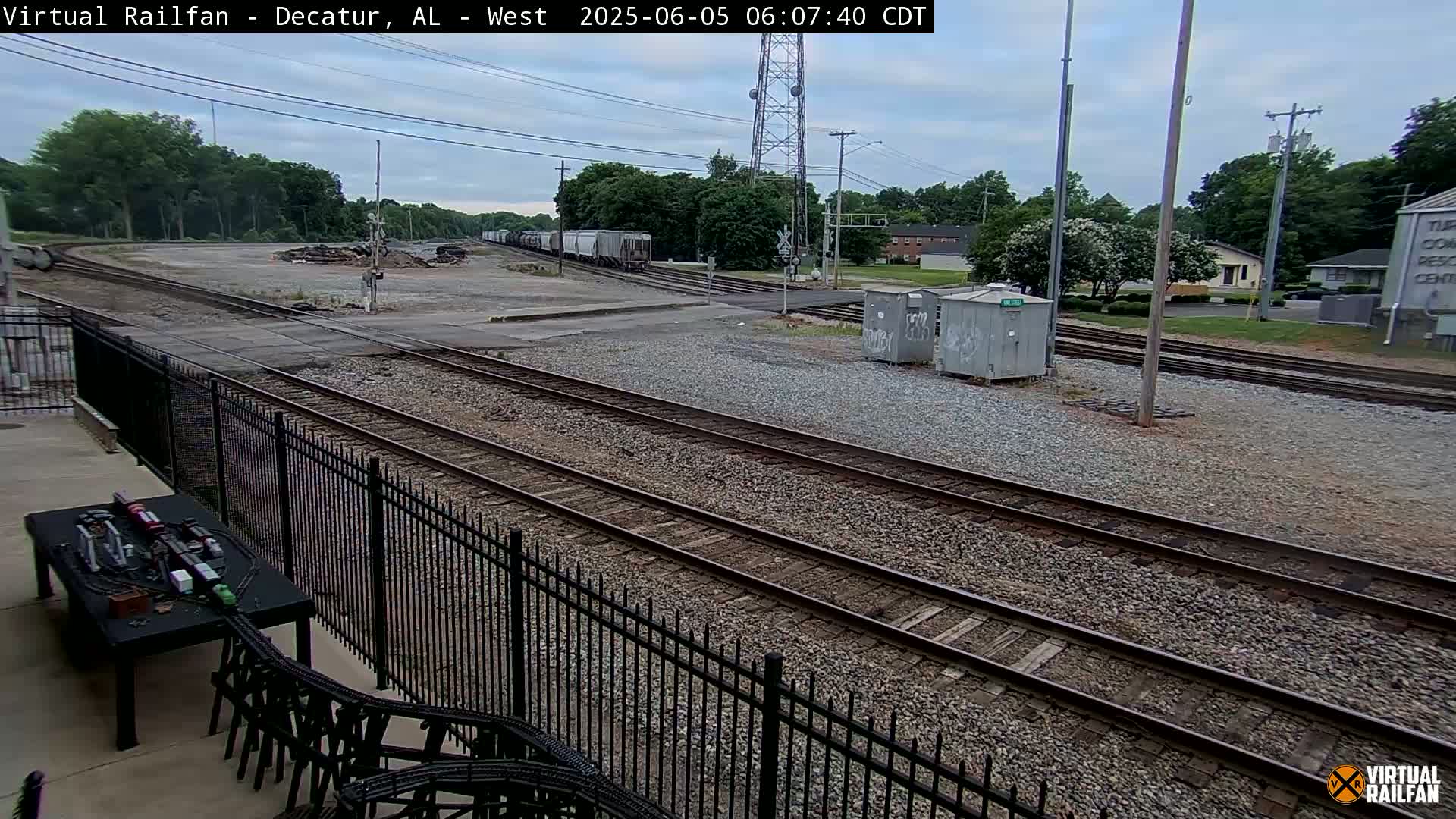 A mostly overcast outdoor view of railroad tracks, a train in the distance, and a model train set on a table near a black metal fence.