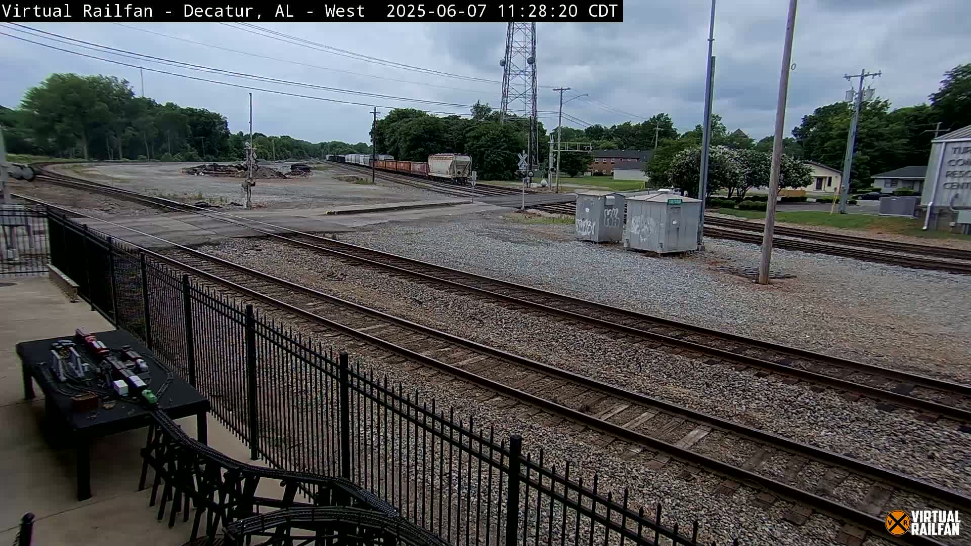 A view of railroad tracks and a train in the distance, under a cloudy sky, from behind a black metal fence.