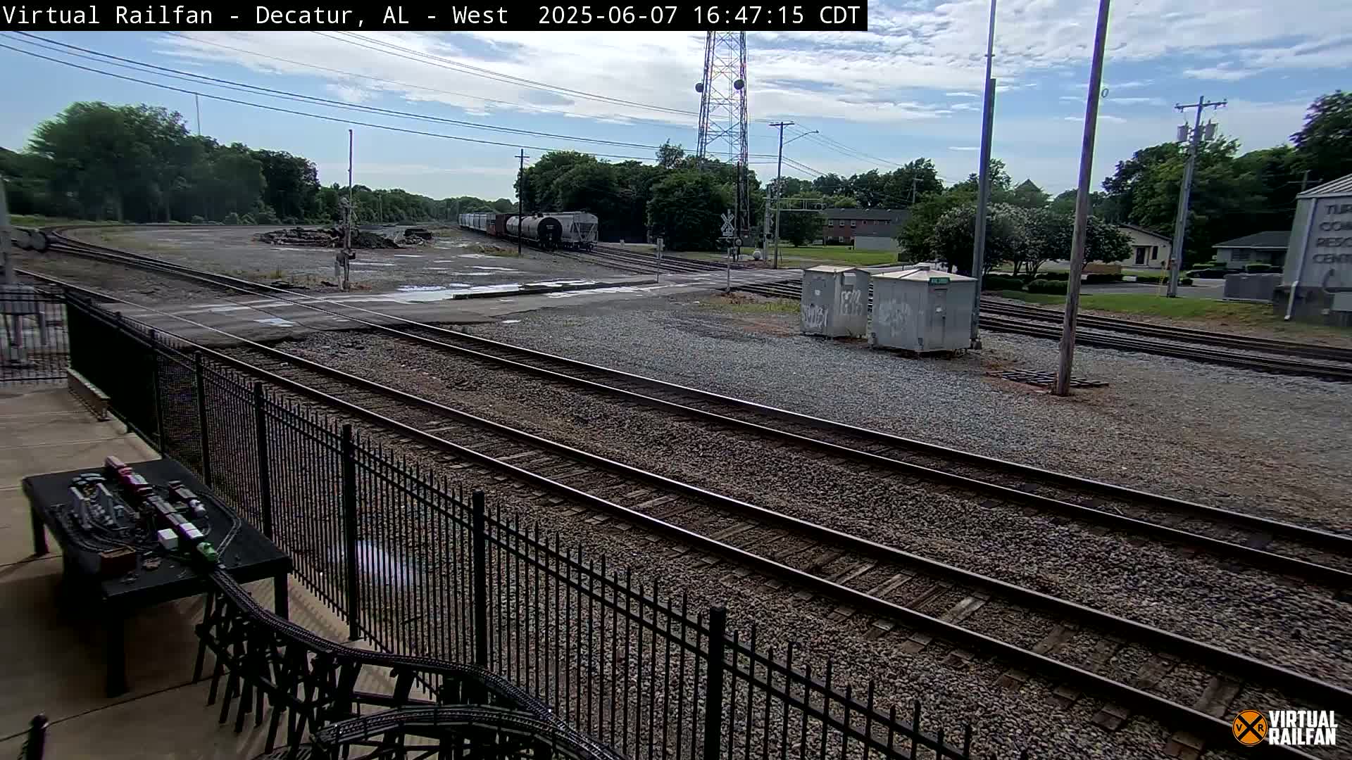 A partially cloudy sky overlooks a railroad yard with several sets of tracks, a train in the distance, and a model train set on a table in the foreground.