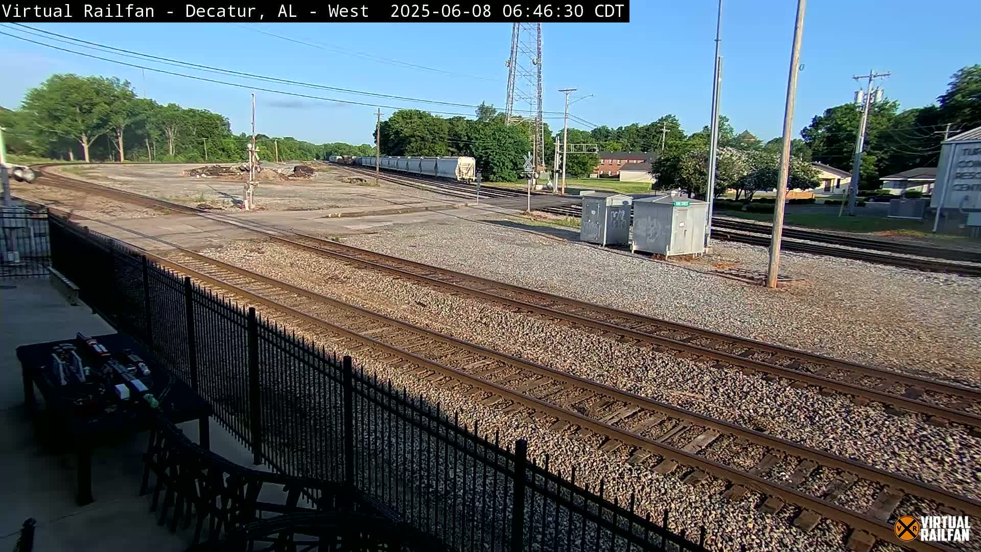 A sunny day shows a view of several railway tracks, a train in the distance, and some buildings and trees in the background, all behind a black metal fence.