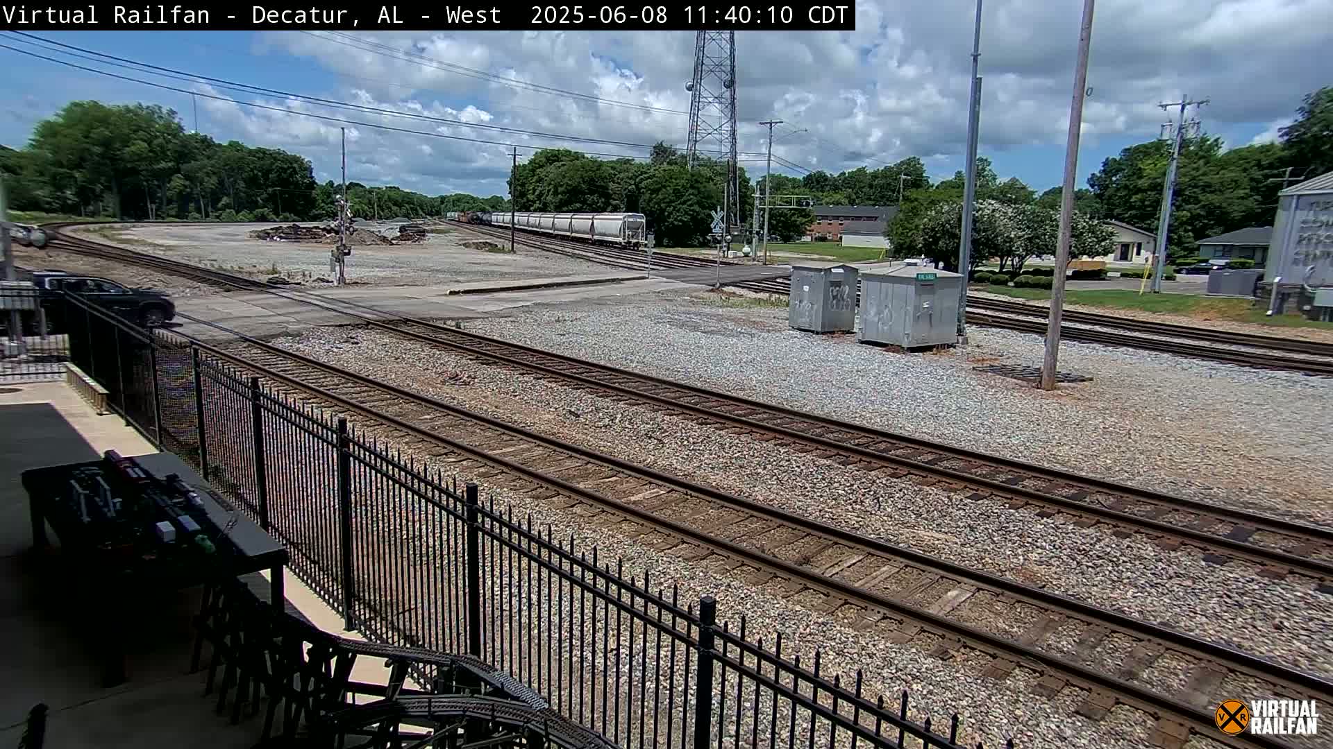 A partly cloudy day shows a train of boxcars passing through a railway intersection near a residential area with trees and a black metal fence in the foreground.