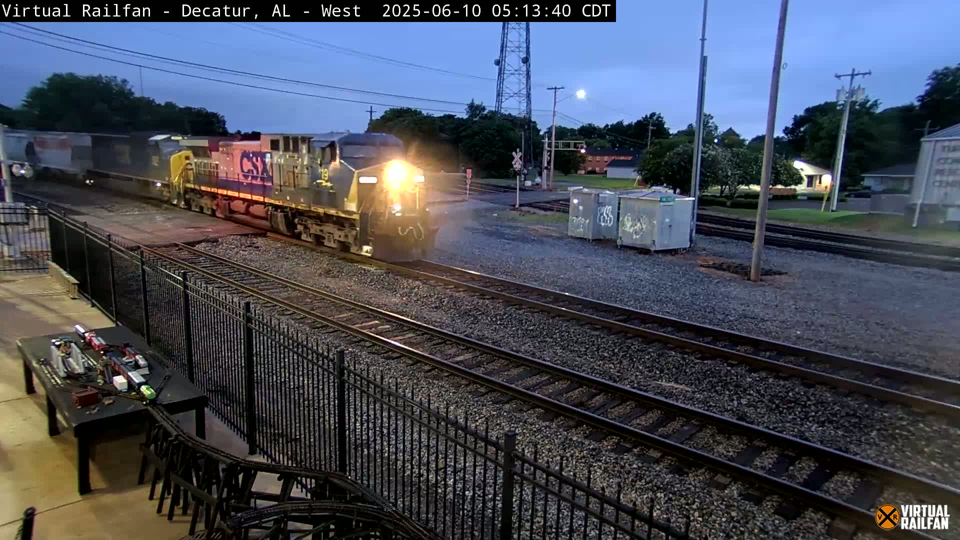 A freight train passes by under a twilight sky, next to a metal fence and model train set.