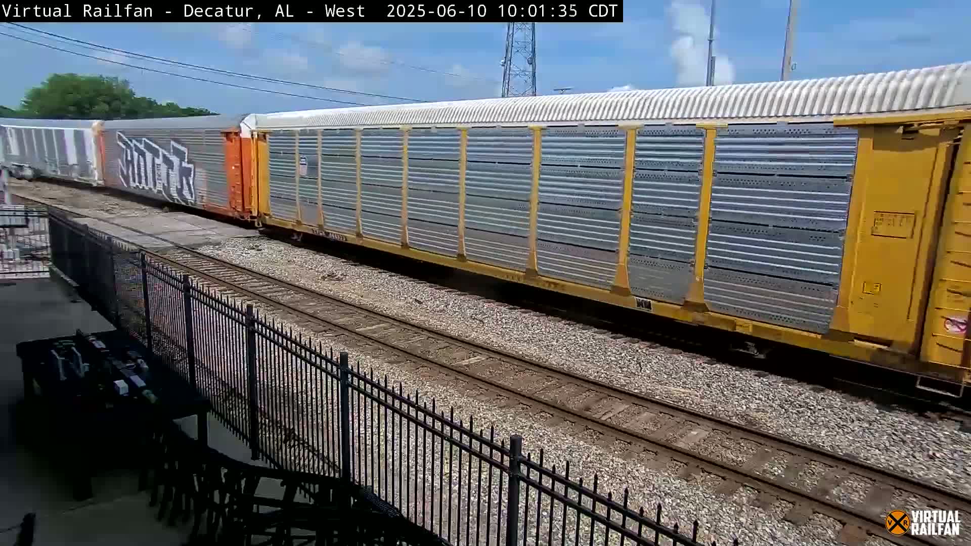 A long freight train with graffiti on some cars passes by a black metal fence on a sunny day.