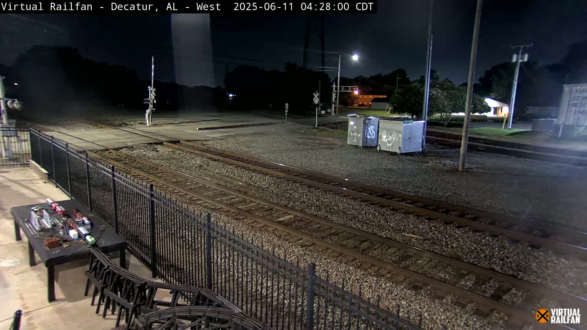 A nighttime view of railroad tracks and a model train set next to a black metal fence under a dark sky.