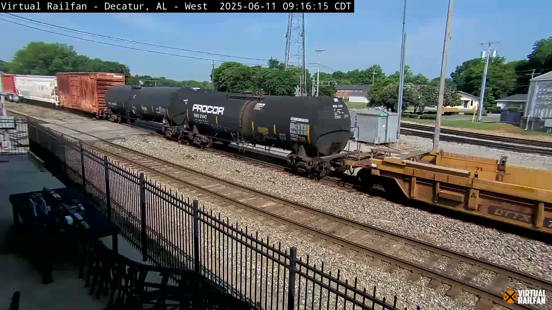 A freight train, consisting of various cars including tank cars and boxcars, moves along railroad tracks on a sunny day.