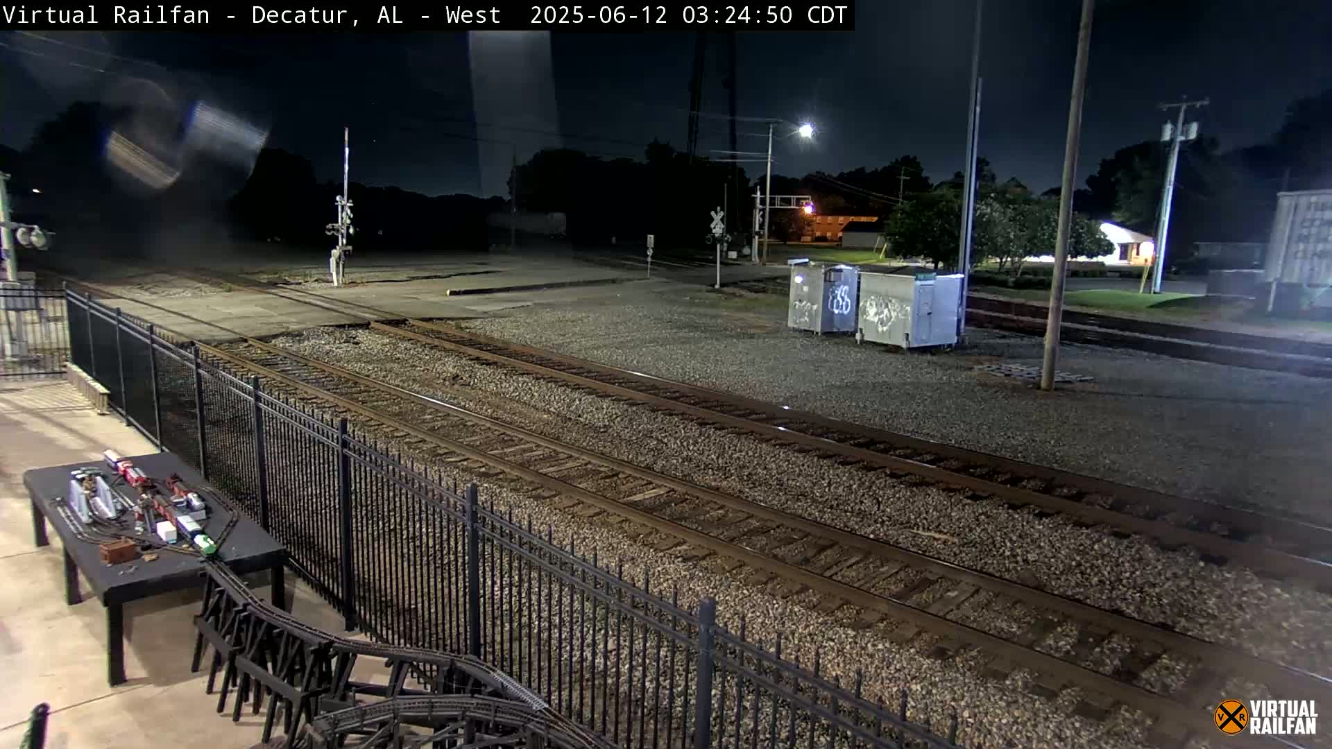 At night, a model train sits on a table next to a black metal fence overlooking train tracks.