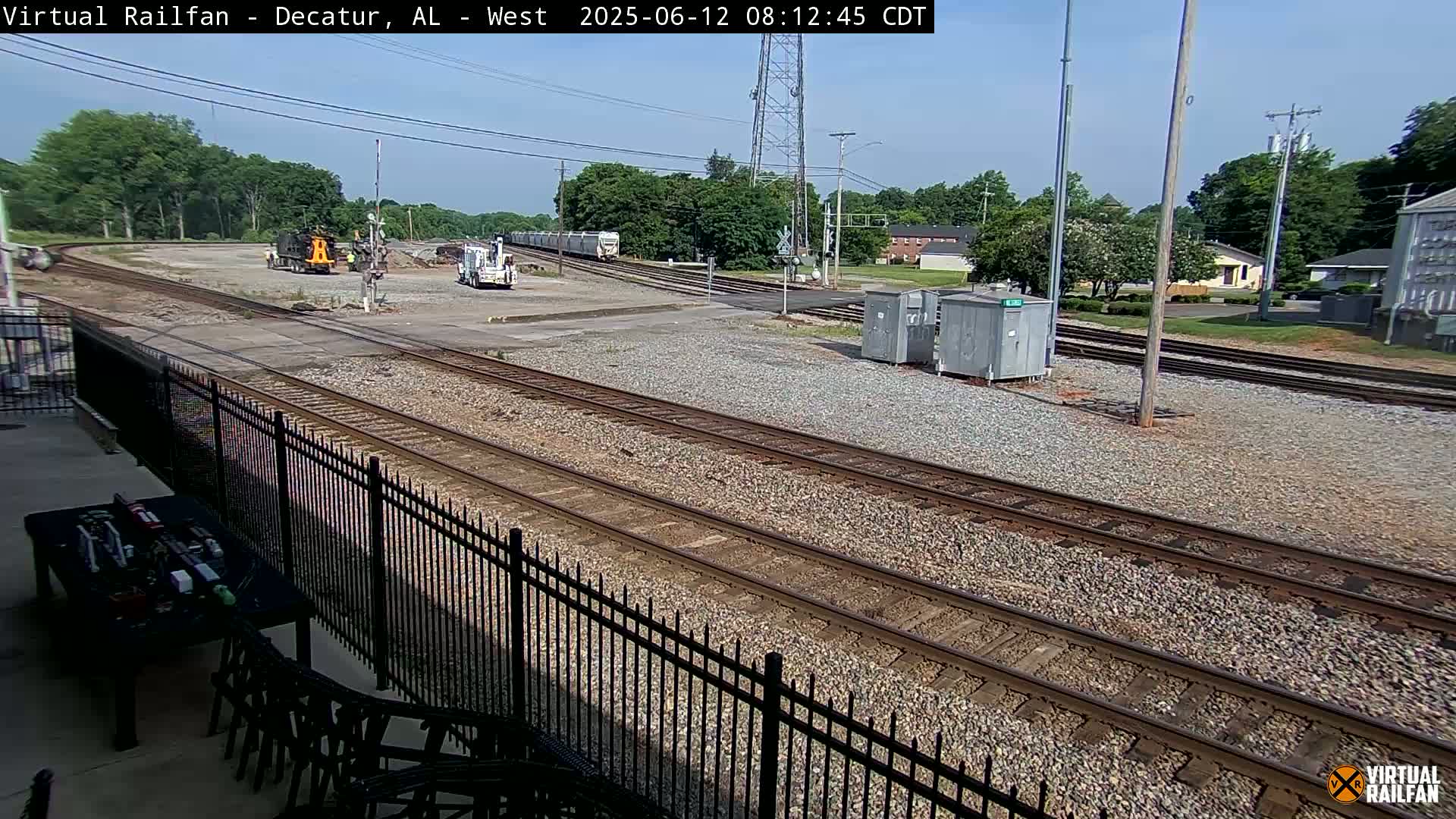 A sunny day reveals several train tracks converging near a railway yard with some rail cars and machinery present, viewed from behind a black metal fence.