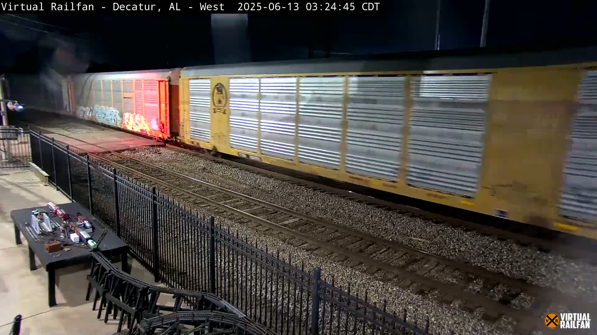 At night, a long freight train passes by a black metal fence, near a model train set on a table.