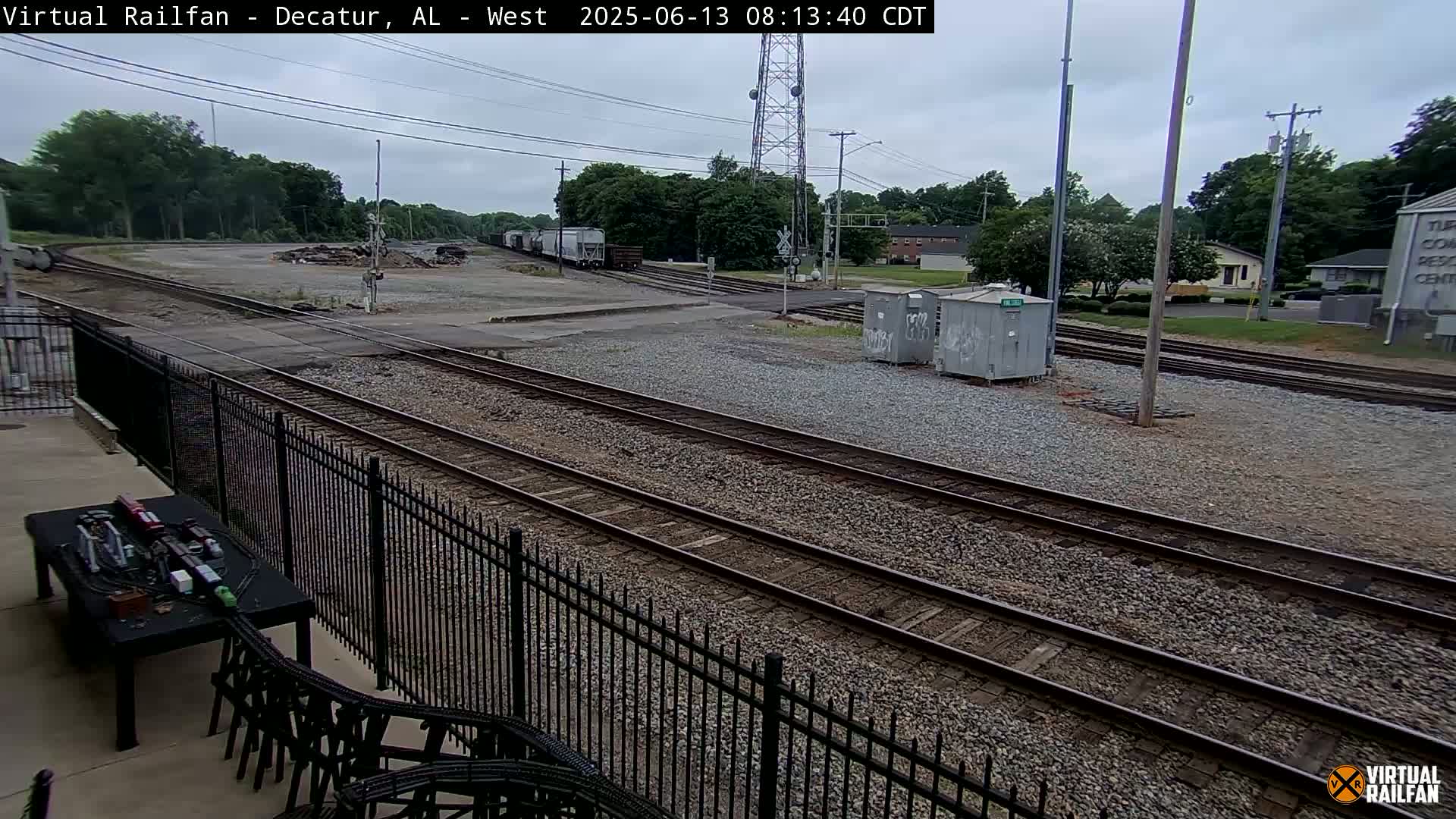 A view of railroad tracks and switching yards under an overcast sky, with some rail cars visible in the distance, and a model train set on a table in the foreground.