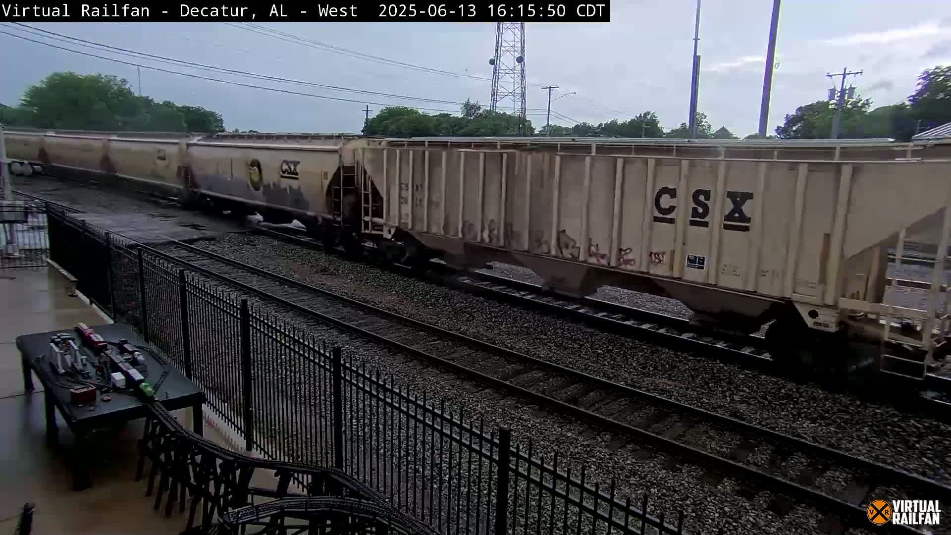 A long freight train passes by on a rainy day, viewed from behind a black metal fence and a patio table.