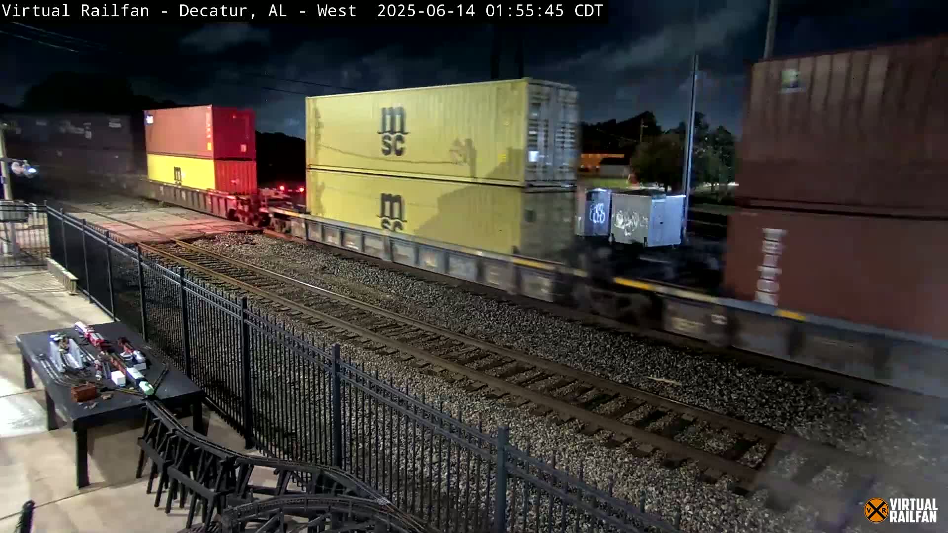 A long freight train passes by on a rainy day, viewed from behind a black metal fence and a patio table.