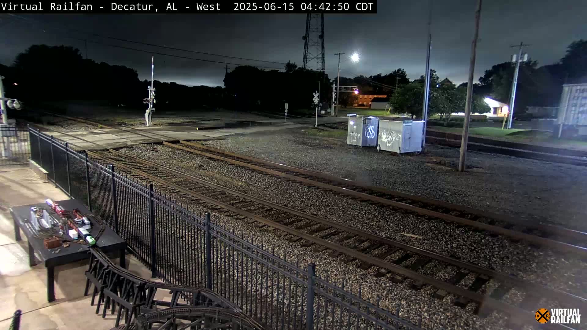 Under a dark, night sky, multiple train tracks stretch across a gravel area, bordered by a black metal fence, with a small model train set visible in the bottom left corner.