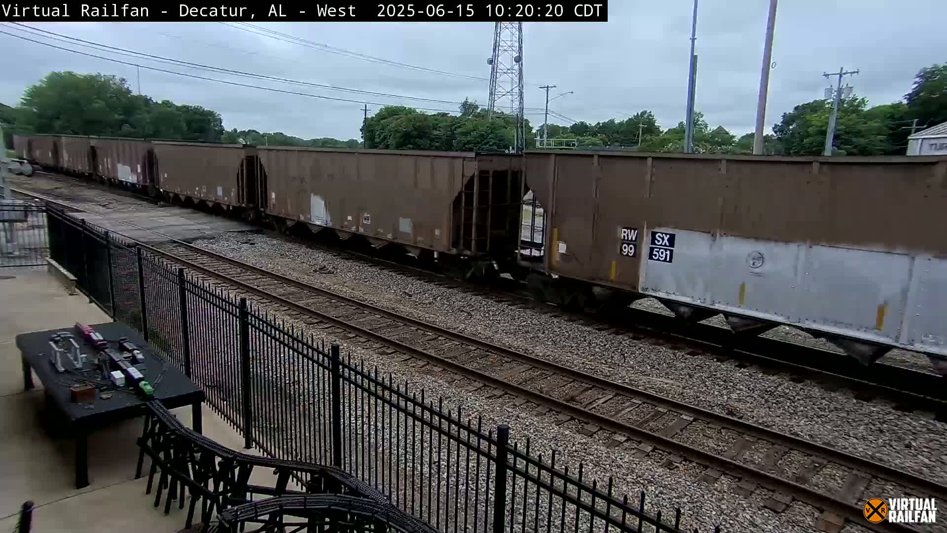 A long freight train passes by on a railway track under an overcast sky, viewed from behind a black metal fence.
