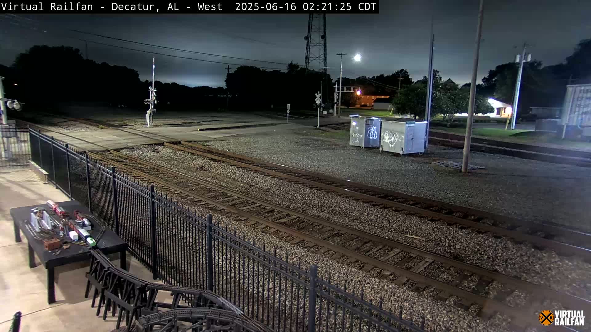 A nighttime view of train tracks, a black metal fence, and a model train set on a table, under a dark, cloudy sky.