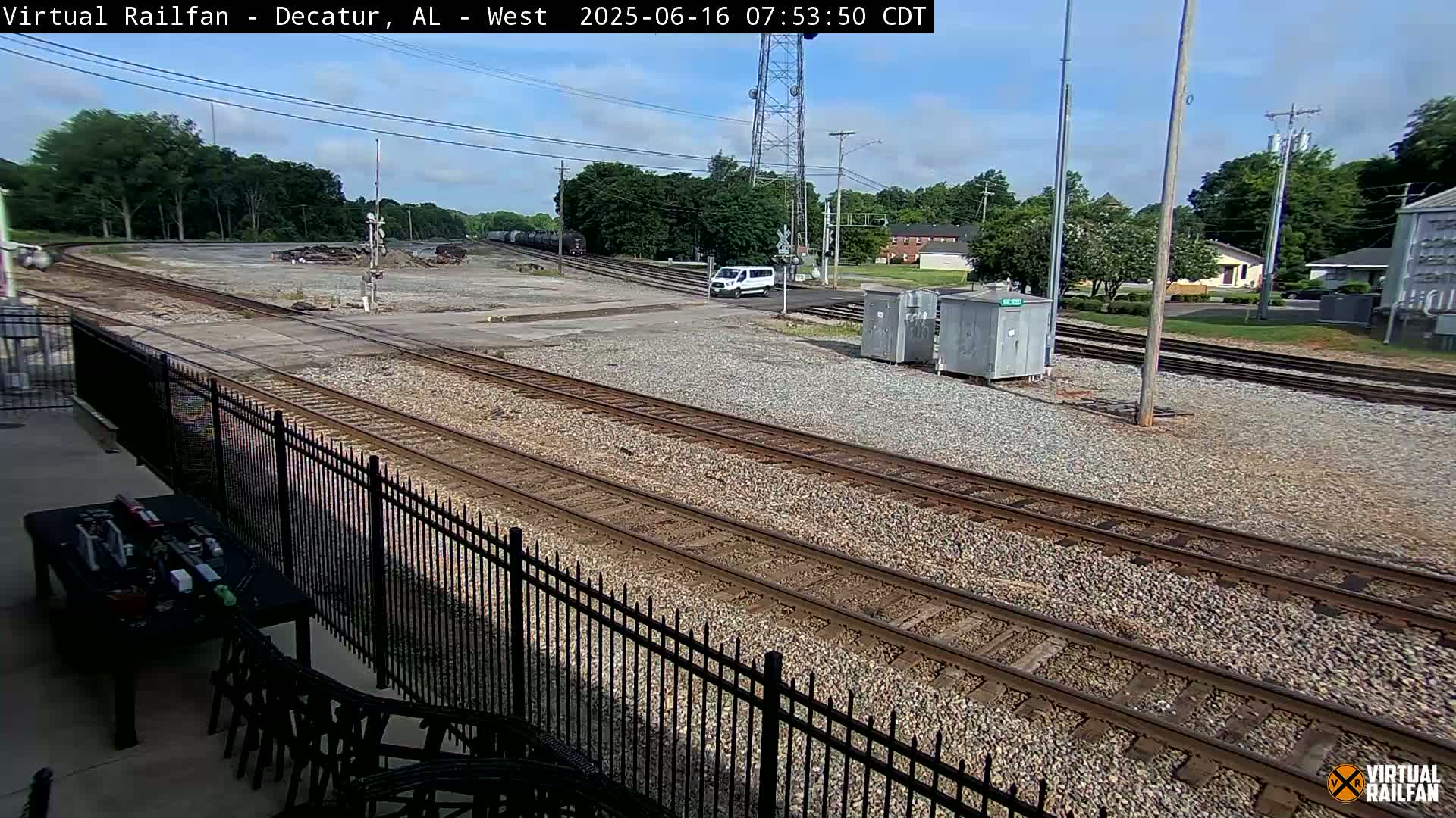 A sunny day reveals a railroad yard with multiple train tracks, a white van, and some small buildings, all bordered by a black metal fence.