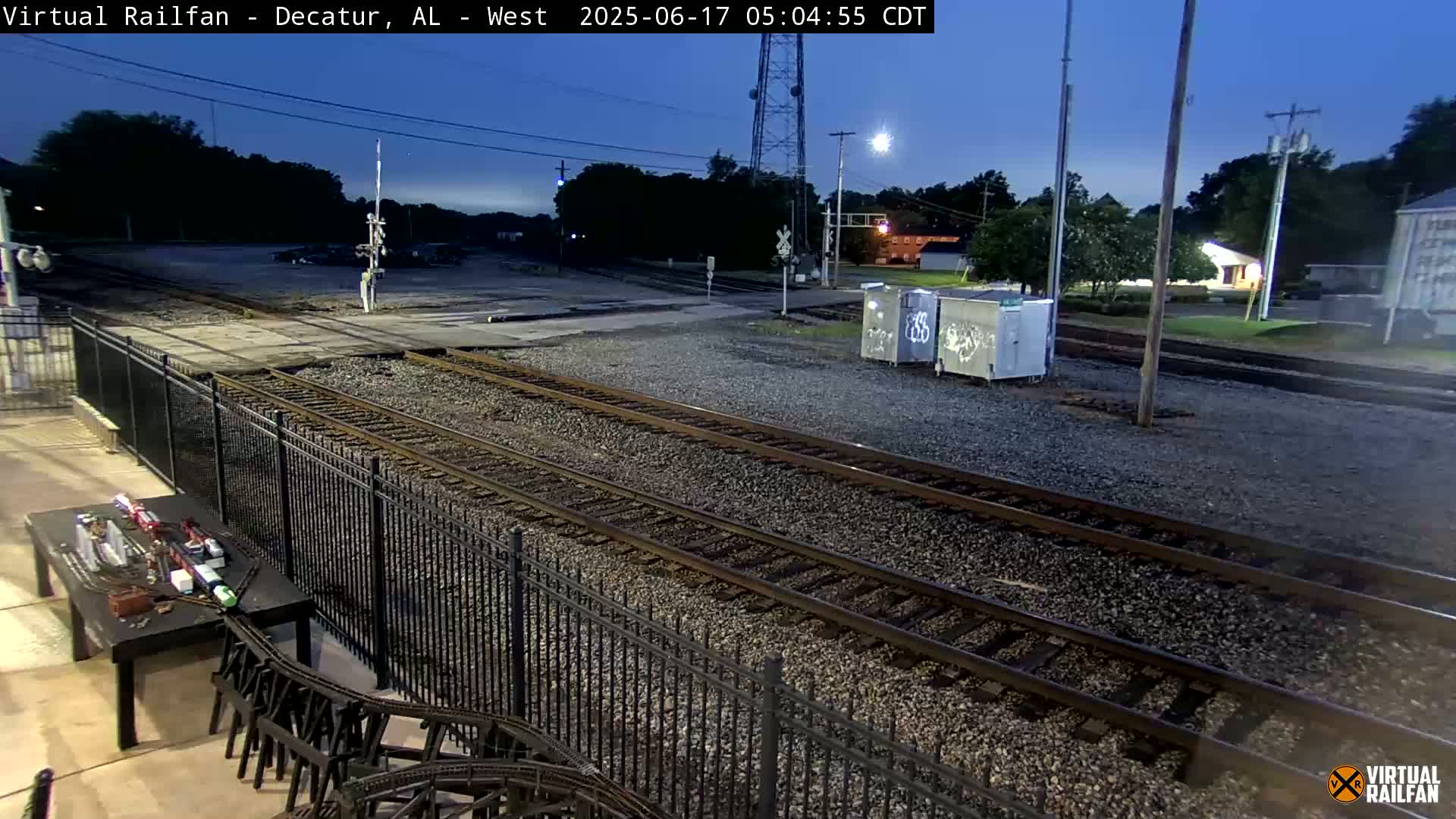 Under a twilight sky, two sets of train tracks are seen extending into the distance, bordered by a black metal fence and a small model train display.