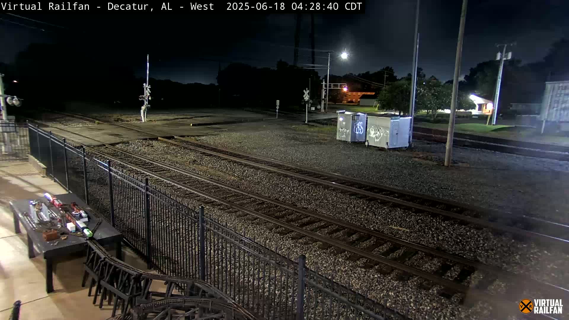 A nighttime view of train tracks, a metal fence, and a table with a model train set, under clear skies.
