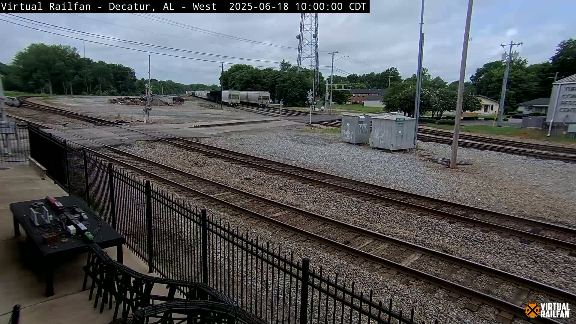 A train of boxcars sits in a rail yard on an overcast day, with multiple sets of train tracks extending into the distance and a black metal fence in the foreground.