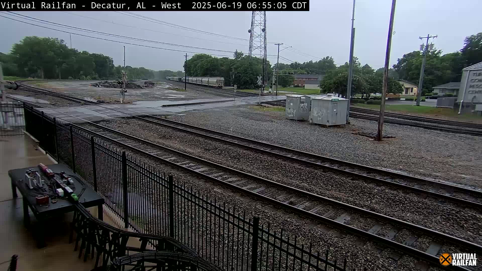 A freight train passes through a railway yard on a rainy day, with several tracks visible and a black metal fence in the foreground.
