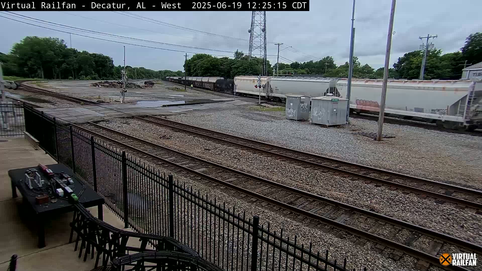 A long freight train passes by a rail yard on an overcast day.