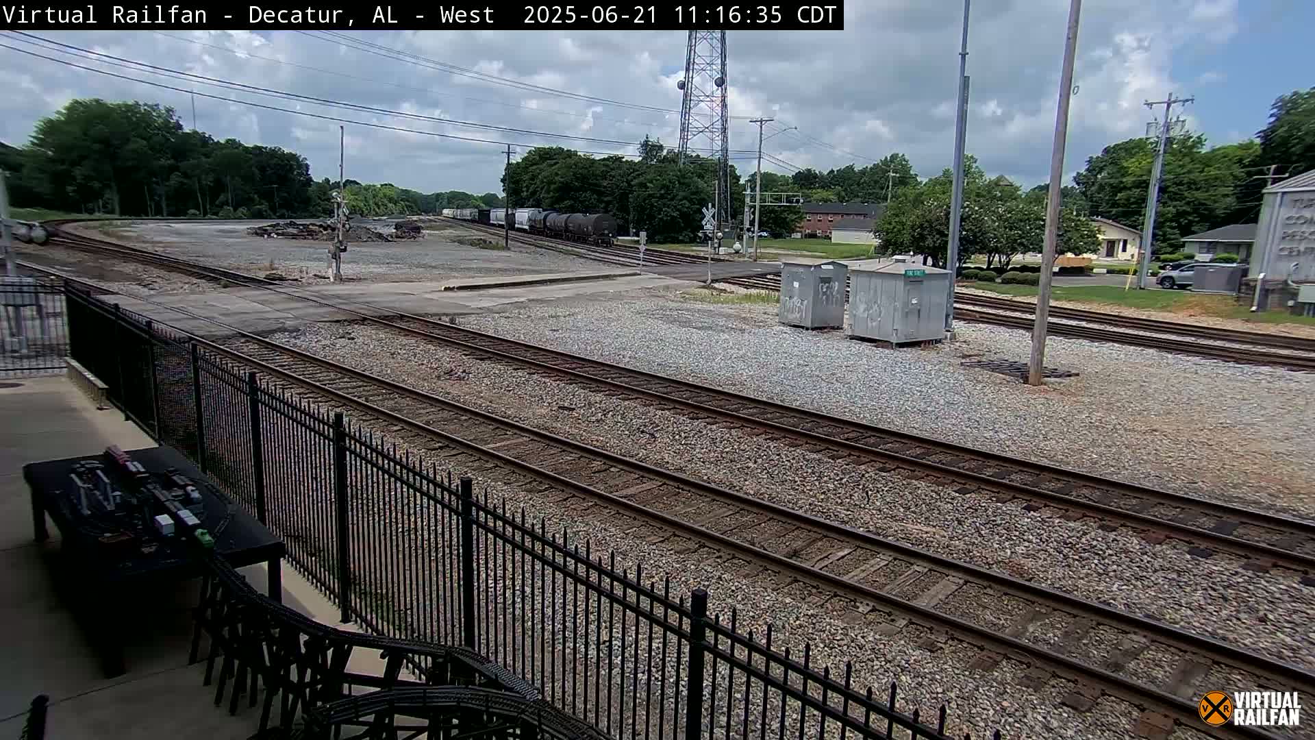 A partly cloudy day shows several railroad tracks converging near a road, with a train of boxcars visible in the distance and a black metal fence in the foreground.