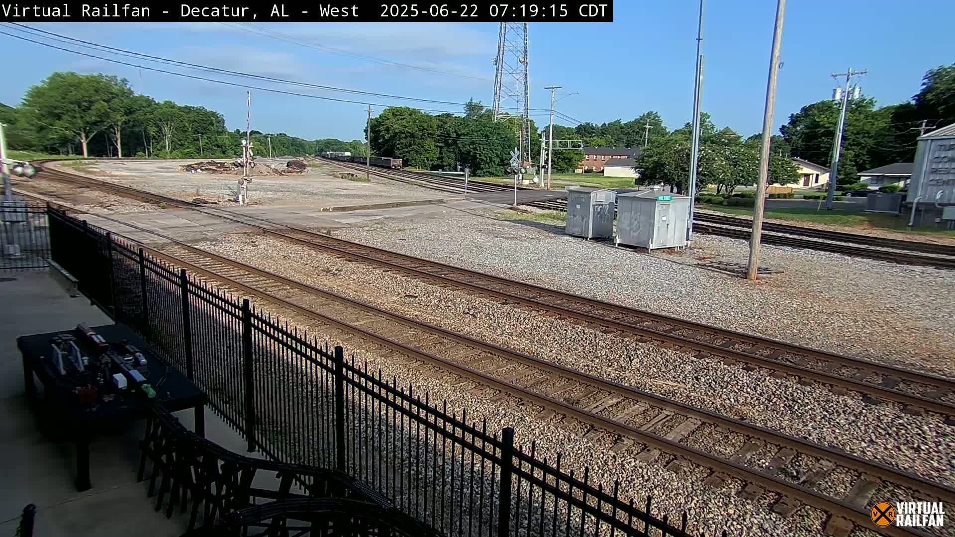 Several sets of train tracks curve through a gravel yard under a clear, sunny sky, next to a black metal fence and some small buildings.
