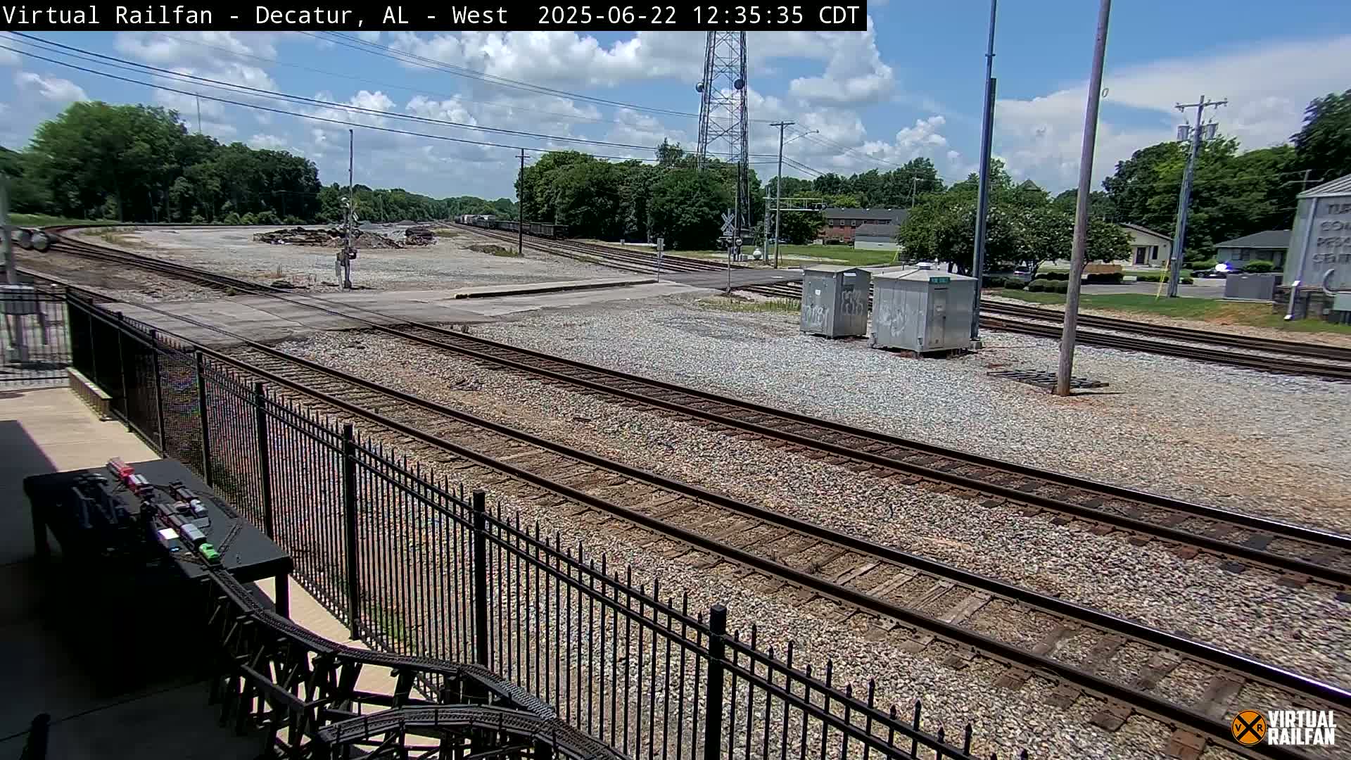 A sunny day shows a railway yard with multiple train tracks, a fence, and some small buildings, with trees and a road visible in the background.