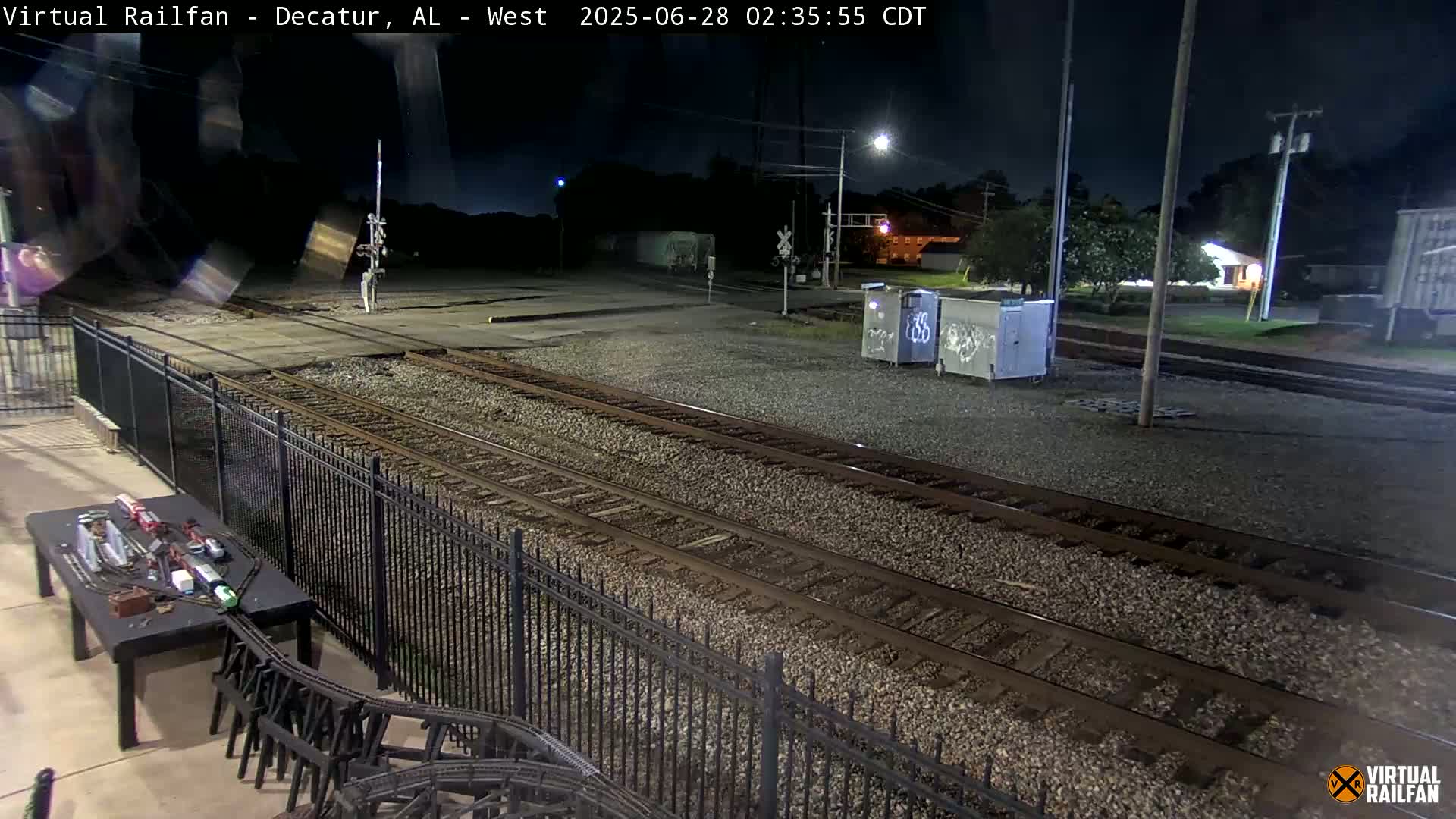 A nighttime view of railroad tracks, bordered by a fence, with a model train set visible on a table in the foreground.