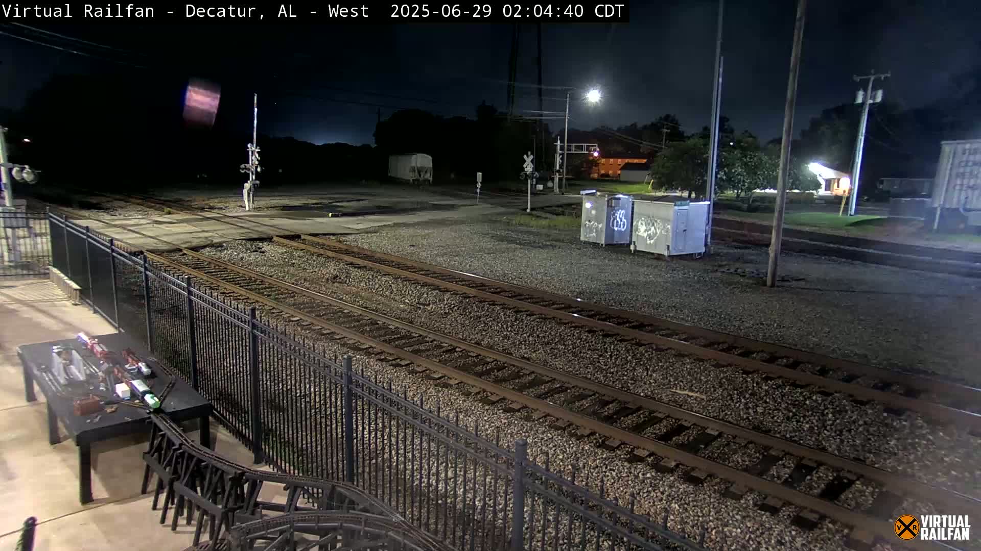A nighttime view of several train tracks, a metal fence, and some graffitied containers under a dark sky.