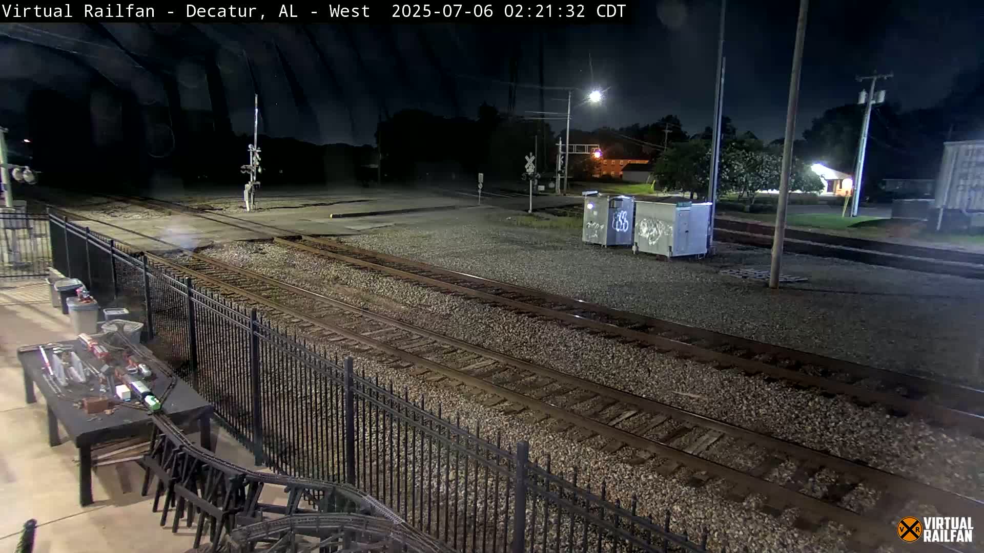 A nighttime view of a train yard with multiple tracks, a metal fence, and a small model train set on a table.