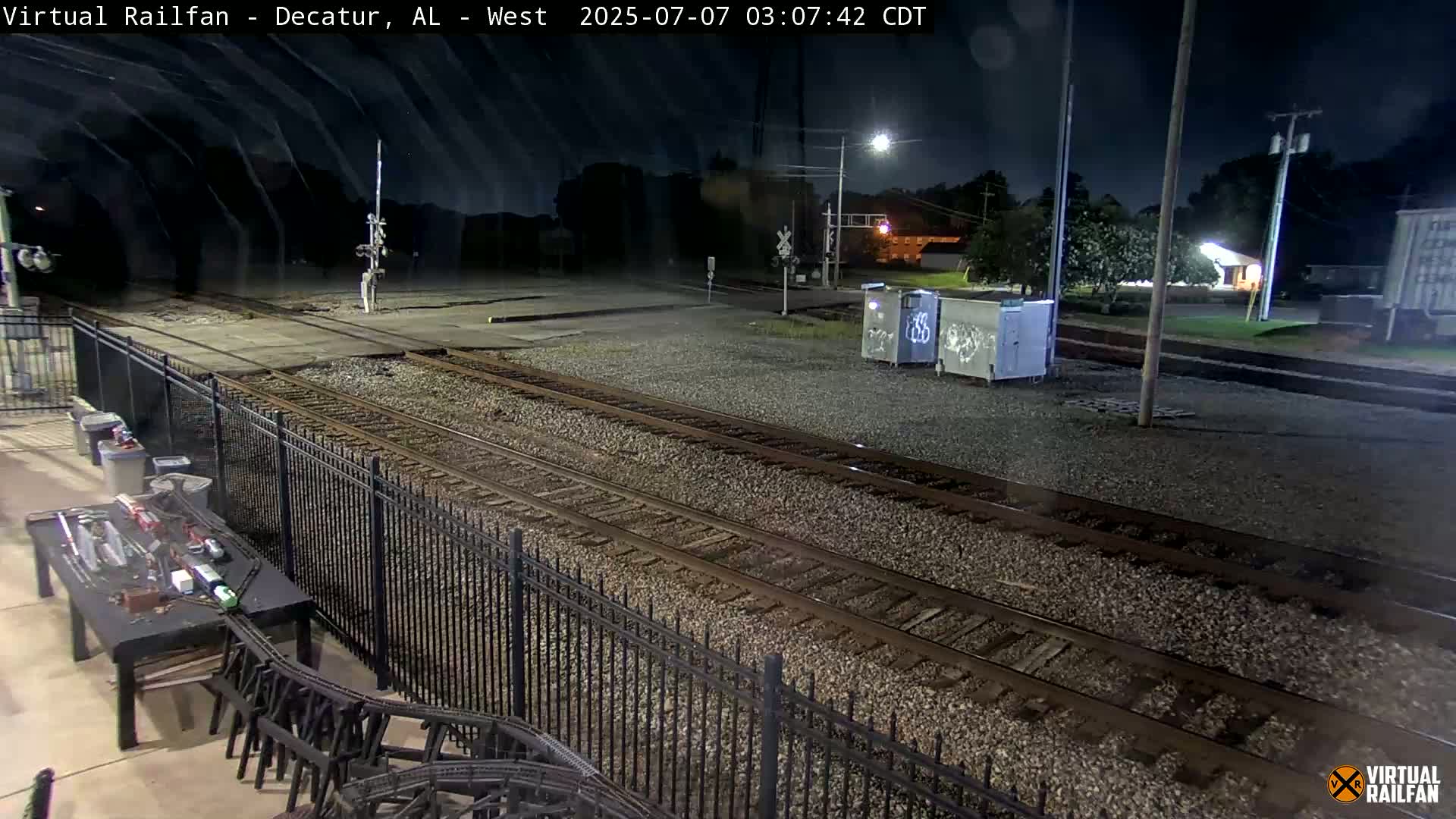 A nighttime view of multiple train tracks bordered by a black metal fence, with a model train set visible on a table in the foreground.