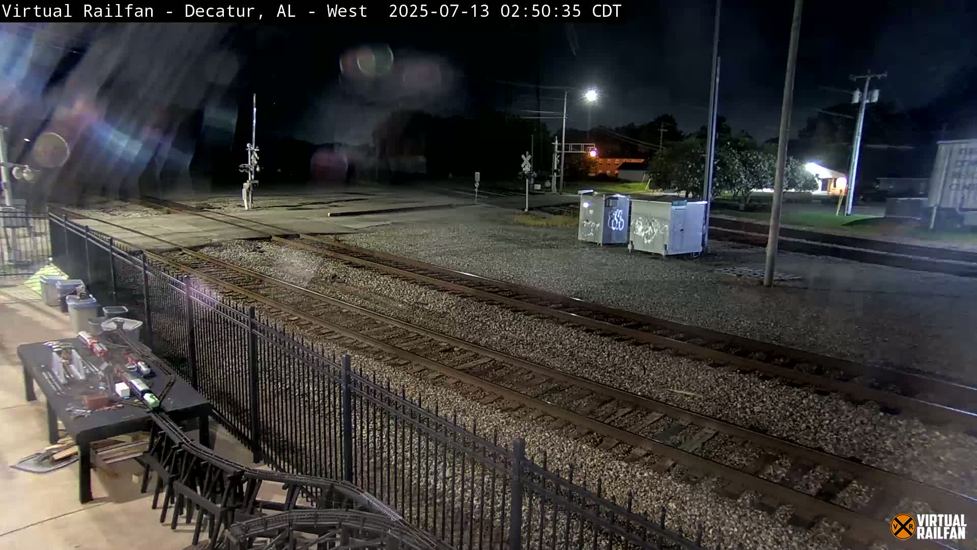 A nighttime view of a train yard with two sets of train tracks, a black metal fence, and several dumpsters.