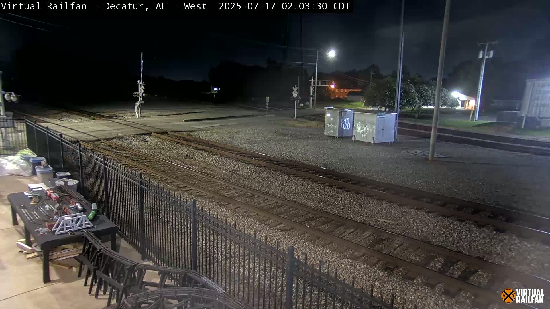 A nighttime view of a railroad crossing with several train tracks, a black metal fence, and a model train setup on a table in the foreground.