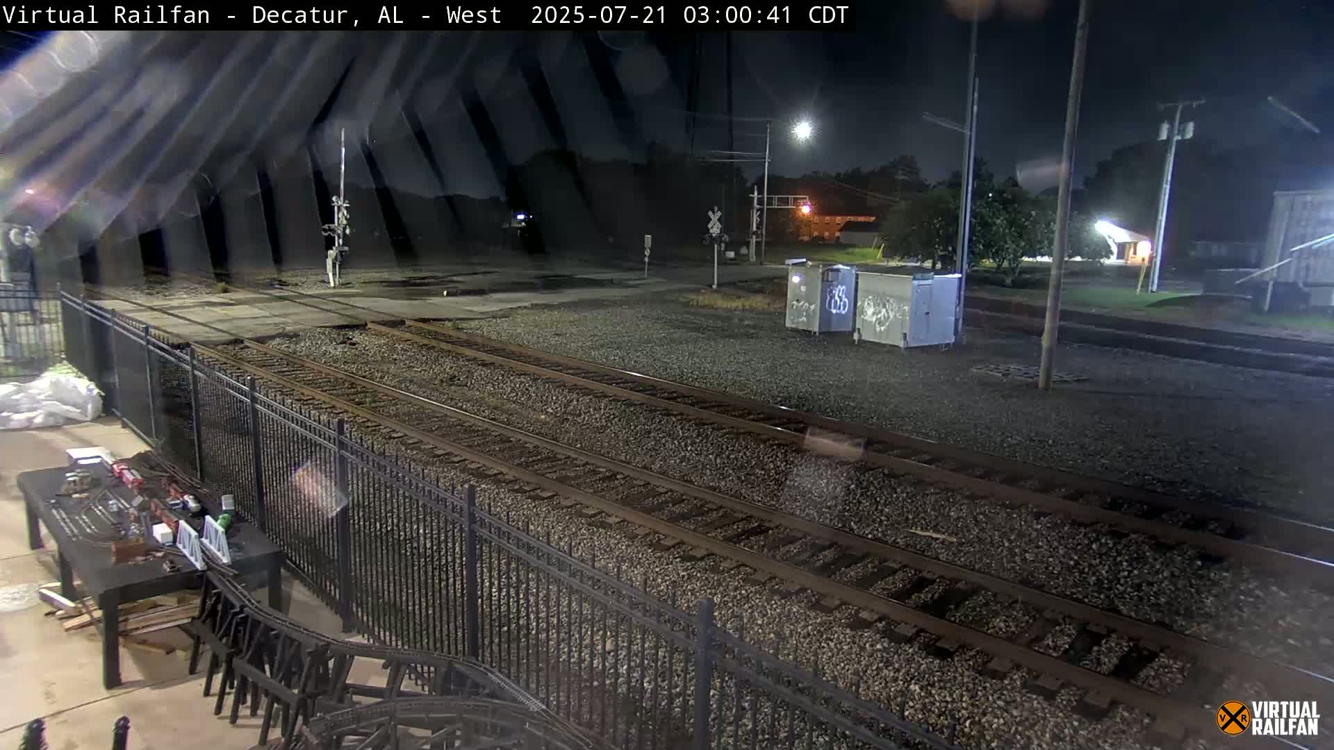 At night, a nighttime view shows train tracks extending into the distance, bordered by a black metal fence, with some small structures and vegetation visible in the background.