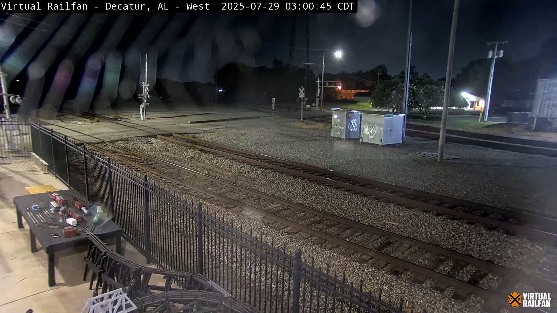 A nighttime view of railroad tracks, bordered by a black metal fence, with several containers nearby and a model train set on a table in the foreground.
