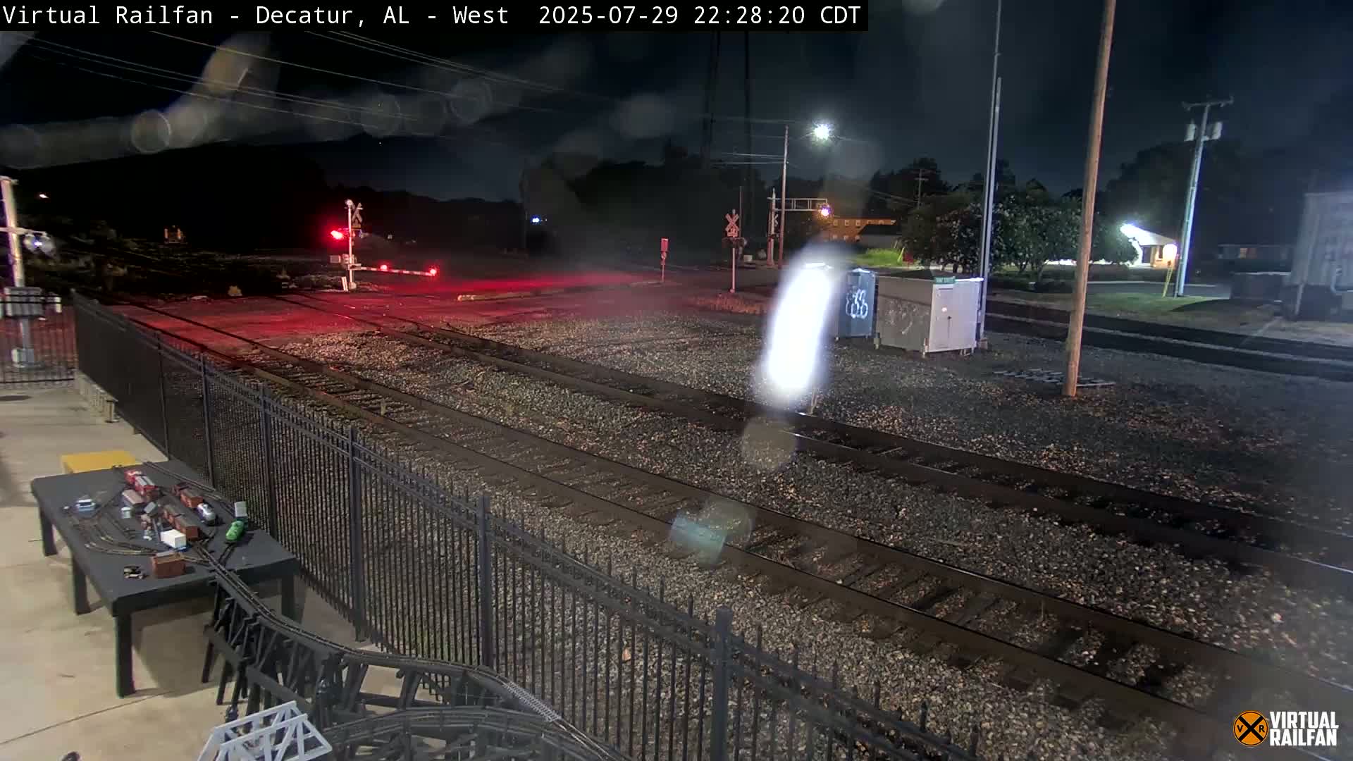 A nighttime view of railroad tracks, bordered by a black metal fence, with several containers nearby and a model train set on a table in the foreground.