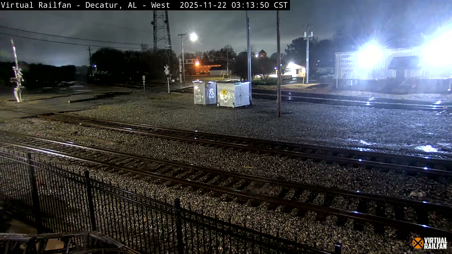A nighttime scene depicts multiple train tracks converging at a railroad crossing in an industrial area, with wet ground reflecting bright artificial lights under a cloudy sky.