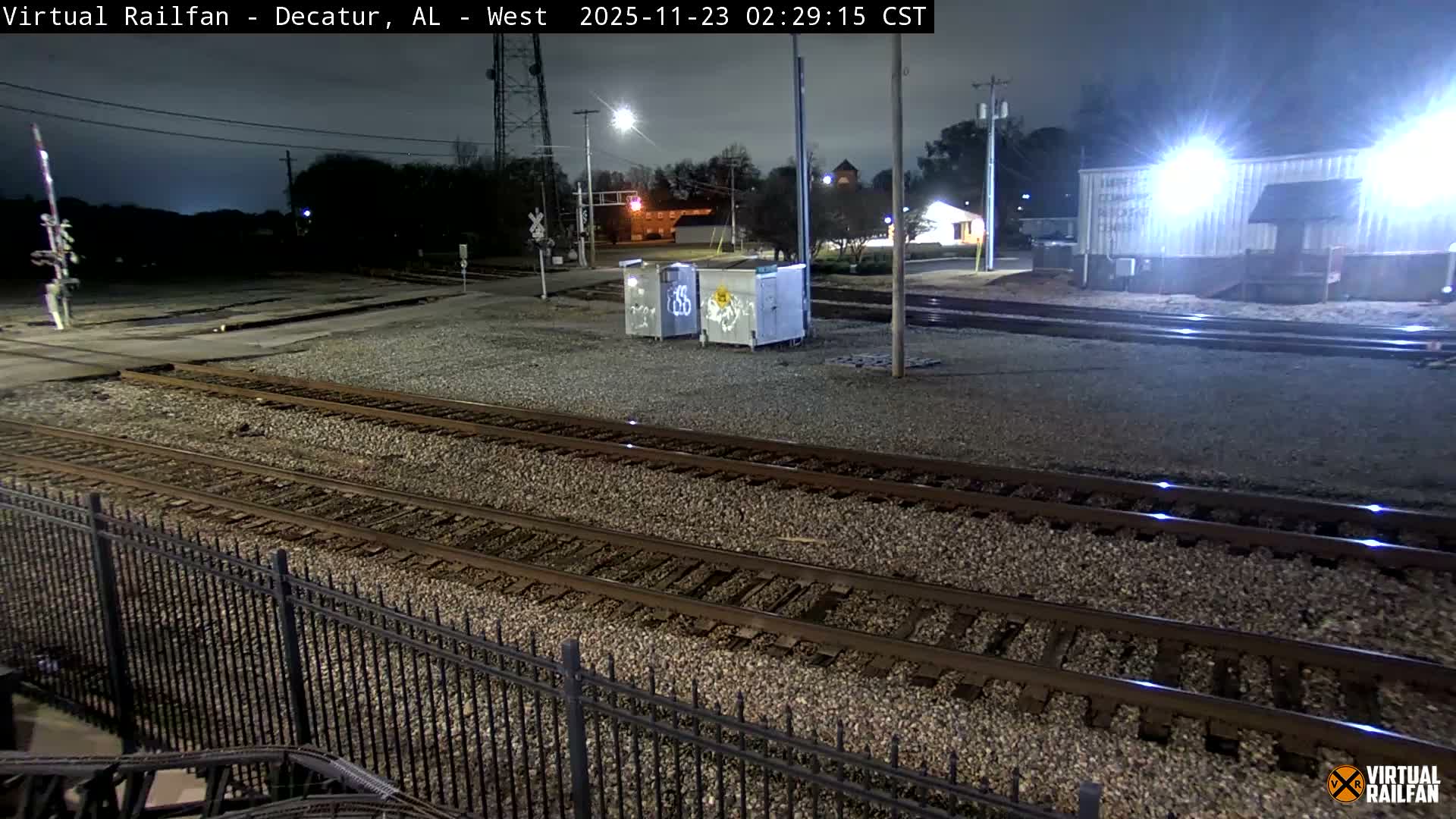 On a dark and cloudy night with dry conditions, multiple parallel railroad tracks extend across the foreground, leading towards a railroad crossing with a signal arm and various illuminated industrial buildings and utility boxes in the midground.