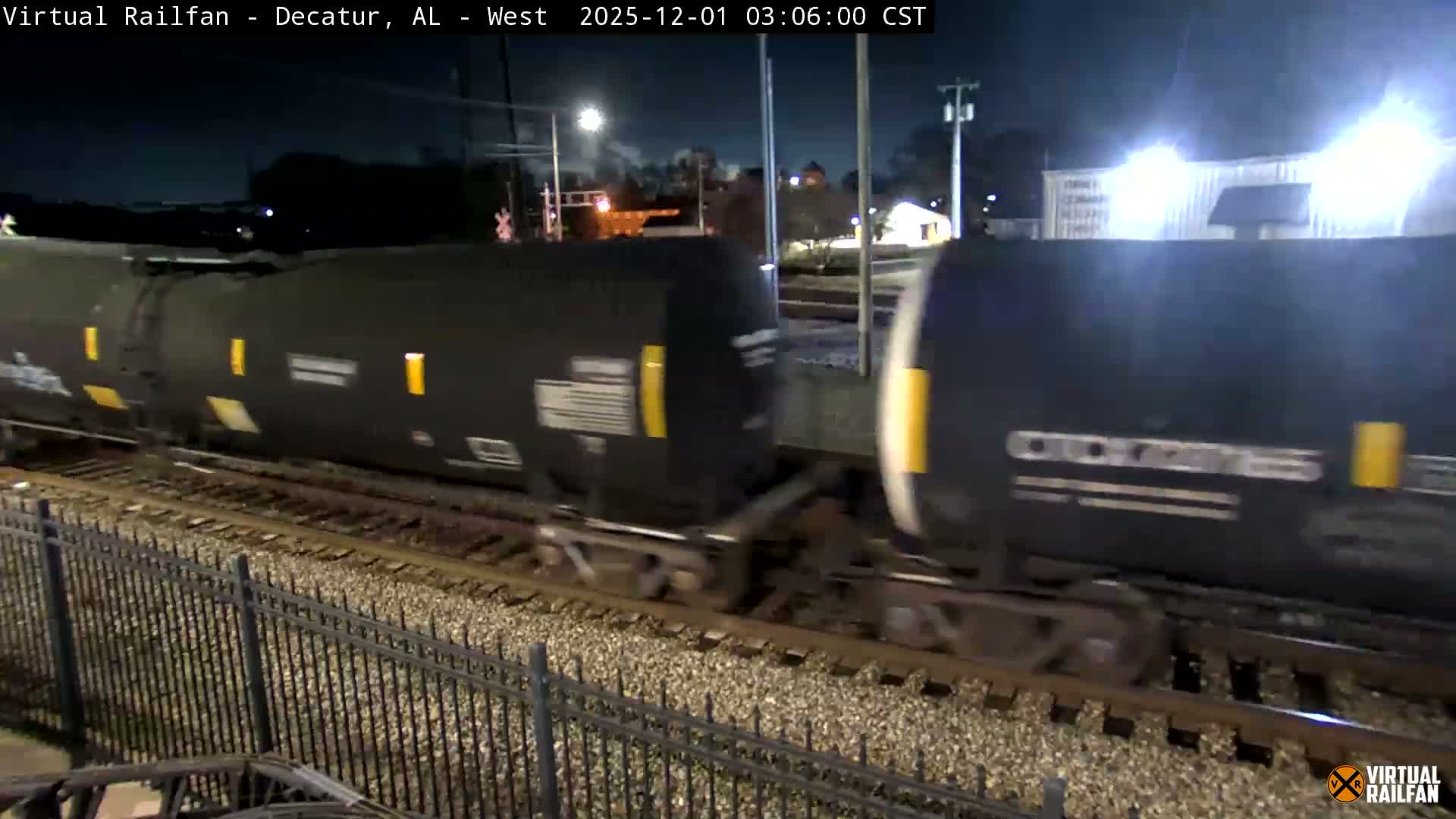 A freight train with black tank cars and yellow markings moves along tracks at night, passing a metal fence in the foreground with illuminated buildings and streetlights visible in the background under a clear sky.
